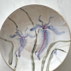 Puede incluir: Un cuenco de cerámica de color blanco roto con un diseño pintado a mano de dos flores azules y moradas similares a los lirios, con tallos y hojas verdes largos y delgados. La superficie interior del cuenco tiene un acabado liso y moteado.