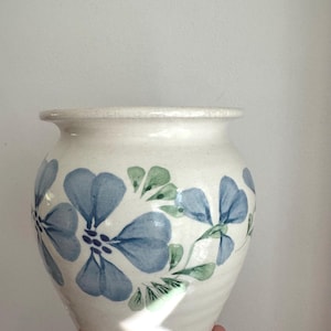 Puede incluir: Un jarrón de cerámica de color crema con cuerpo redondeado y borde acampanado. El exterior presenta un patrón floral pintado a mano que consiste en pétalos azules y hojas verdes dispuestos en una banda horizontal alrededor del centro.