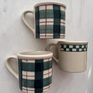 Puede incluir: Tres tazas de cerámica con asas. Dos tazas presentan un patrón de cuadros verdes y blancos, mientras que la tercera tiene una base color crema con un borde a cuadros verdes. Todas las tazas tienen un borde y un asa color crema.