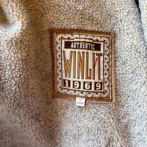 Pode incluir: Close-up de uma etiqueta castanha e bege com o texto "AUTHENTIC WINLIT 1969" sobre um tecido bege texturizado. A etiqueta &eacute; retangular com uma borda decorativa e uma pequena etiqueta que diz "MADE IN CHINA".
