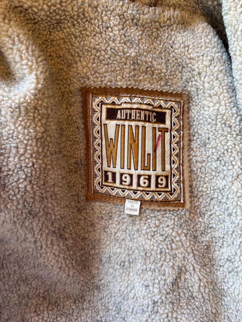 Pode incluir: Close-up de uma etiqueta castanha e bege com o texto "AUTHENTIC WINLIT 1969" sobre um tecido bege texturizado. A etiqueta &eacute; retangular com uma borda decorativa e uma pequena etiqueta que diz "MADE IN CHINA".