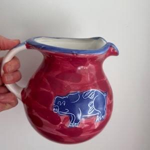 Puede incluir: Una jarra de cerámica con un diseño veteado rojo y blanco. La jarra tiene una ilustración de cerdo azul en la parte delantera y un asa blanca. El borde está ribeteado en azul. La jarra está sujeta por una mano.