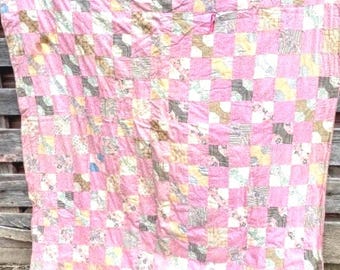 May include: A patchwork quilt featuring a repeating grid pattern of solid pink fabric squares alternating with squares of various patterned fabrics in shades of brown, white, and muted yellow, creating a structured and colorful textile design.