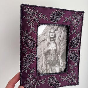 Puede incluir: Un marco de fotos rectangular, de color ciruela, con intrincados bordados florales en plata y negro. El marco está adornado con pequeñas cuentas oscuras en los bordes. Contiene una fotografía en blanco y negro de una mujer.