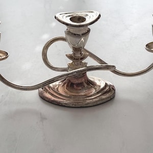 Puede incluir: Candelabro plateado con tres candeleros. El soporte central es triangular y los otros dos son redondeados. Los brazos curvos se extienden desde una base circular. El candelabro es de metal y tiene un aspecto deslustrado.