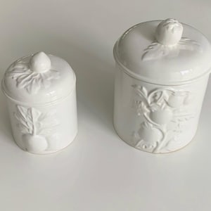 Puede incluir: Dos botes de cerámica blanca con tapas, ambos con diseños florales en relieve. El bote más grande tiene un diseño de tomates en una vid, mientras que el bote más pequeño tiene un diseño de una sola flor.