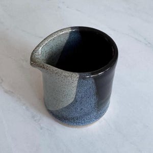 Puede incluir: Una pequeña jarra de cerámica hecha a mano con un pico. La jarra presenta un diseño de bloques de color con secciones de gris moteado, azul texturizado y negro brillante. El interior es negro y la base no está esmaltada.