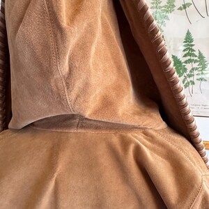Pode incluir: Close-up de um casaco com capuz de camur&ccedil;a castanho-claro. O capuz &eacute; o ponto focal, mostrando a textura macia e os detalhes das costuras. As bordas do capuz apresentam costuras decorativas. O casaco parece ser feito de um material macio semelhante &agrave; camur&ccedil;a.