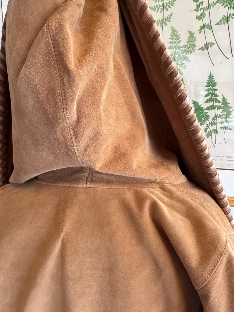 Pode incluir: Close-up de um casaco com capuz de camur&ccedil;a castanho-claro. O capuz &eacute; o ponto focal, mostrando a textura macia e os detalhes das costuras. As bordas do capuz apresentam costuras decorativas. O casaco parece ser feito de um material macio semelhante &agrave; camur&ccedil;a.