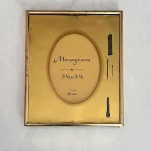 May include: A square, gold-colored picture frame with an oval opening. The frame has the word "Monogram" in script, with dimensions of 3.75 x 4.75 inches. The frame is on a white surface.