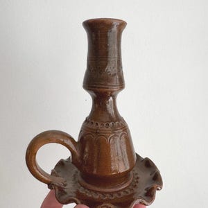Puede incluir: Un candelero de cerámica marrón con asa y base festoneada. El candelero tiene una parte superior alta y cilíndrica y detalles decorativos alrededor de la base y el tallo. El asa es curva y la base es redonda.
