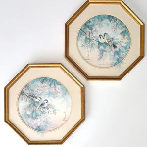 May include: Two framed bird prints. Each print features a circular image of birds perched on flowering branches, with a soft color palette of blue, white, and pink. The frames are octagonal with a gold finish.