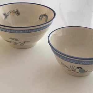 Puede incluir: Dos cuencos de cerámica blanquecina con bordes azules y diseños florales azules decorativos. Los cuencos tienen un diseño sencillo con detalles florales verdes y naranjas pintados. Los cuencos son probablemente para servir alimentos.