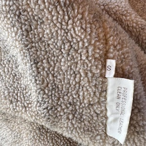 Pode incluir: Close-up de um tecido bege texturizado com uma pequena etiqueta branca. A etiqueta diz "S" e "PROFESSIONAL LEATHER CLEAN ONLY". O tecido tem uma textura densa e irregular, com detalhes cinza e marrom mais escuros.