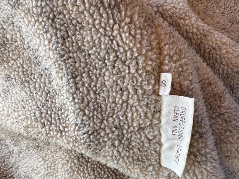 Pode incluir: Close-up de um tecido bege texturizado com uma pequena etiqueta branca. A etiqueta diz "S" e "PROFESSIONAL LEATHER CLEAN ONLY". O tecido tem uma textura densa e irregular, com detalhes cinza e marrom mais escuros.