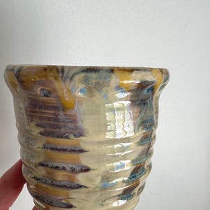 Puede incluir: Una taza de cerámica con borde acampanado y exterior acanalado y texturizado. El esmalte presenta una mezcla de tonos crema, amarillo, marrón y azul, creando una apariencia arremolinada y moteada en la superficie del recipiente brillante.