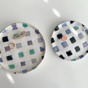 May include: Two ceramic trays with a checkered pattern in shades of blue, green, and black. The larger tray has a ring and a hair clip on it.