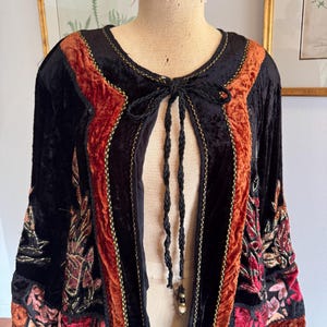May include: Black velvet jacket with rust-colored panels and gold trim. The jacket features floral embroidery on the sleeves and a braided tie closure. The jacket is displayed on a mannequin.