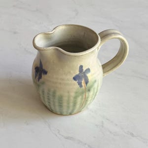 Puede incluir: Una pequeña jarra de cerámica color blanco roto con cuerpo redondeado y una sola asa. El exterior presenta motivos florales azules pintados a mano y pinceladas verdes verticales alrededor de la base, con un acabado esmaltado y liso.