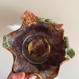 Puede incluir: Un cuenco de cerámica hecho a mano con una forma orgánica única. El cuenco presenta un rico esmalte marrón oscuro con toques de azul y dorado. Adornado con detalles esculpidos, incluyendo una rosa color melocotón, vides verdes y una rana.