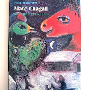 May include: A book cover for "First Impressions Marc Chagall" by Howard Greenfield. The cover features a vibrant, surreal painting with a green bird, a red horse, and a snowy landscape with a small cabin and a person on a sled.