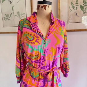 May include: A vintage, long-sleeved dress with a vibrant paisley pattern in pink, orange, purple, and green. The dress features a collared neckline, button-down front, and a matching tie belt. The dress is displayed on a mannequin.