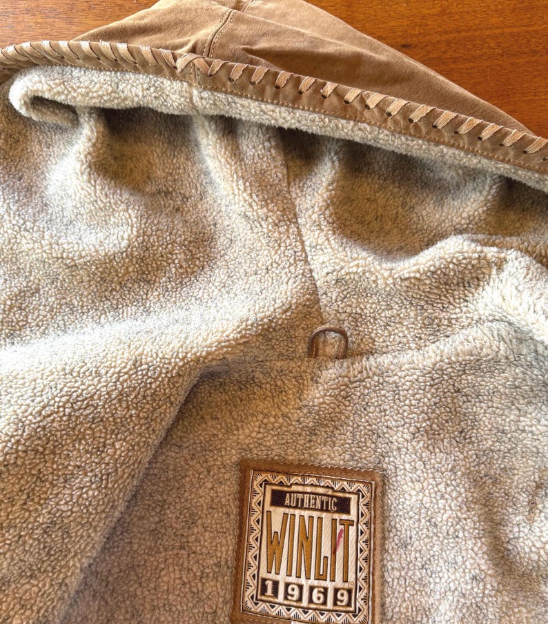 Pode incluir: Close-up de uma jaqueta de camur&ccedil;a marrom com capuz e interior macio e fofo. O capuz &eacute; adornado com costuras decorativas. A jaqueta apresenta uma etiqueta que diz "AUTHENTIC WINLIT 1969".