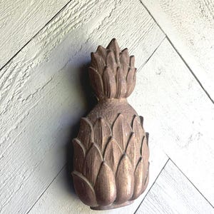 Puede incluir: Un llamador de puerta de piña de color dorado con un diseño detallado. El llamador está montado en una puerta de madera blanca con un patrón de espiga.