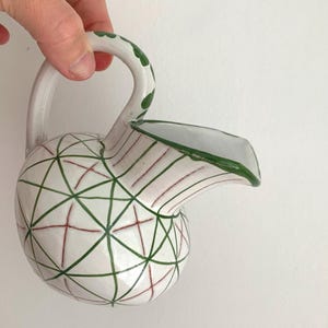 Puede incluir: Una jarra de cerámica blanca con un diseño geométrico verde y rojo. La jarra tiene un cuerpo redondeado y un pico curvo.