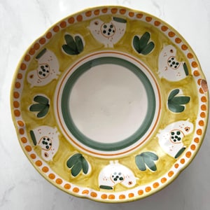 May include: A decorative ceramic plate with a yellow rim and a white center. The plate features a repeating pattern of white birds with black spots and green leaf-like shapes. The rim has orange dots.