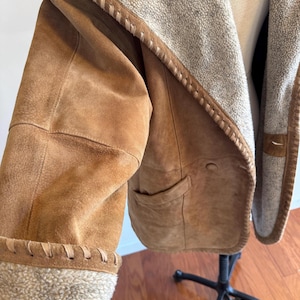 Pode incluir: Casaco de camur&ccedil;a castanho claro com forro felpudo creme e acabamento costurado. O casaco tem capuz e um bolso. As mangas s&atilde;o aparadas com o mesmo material felpudo. O casaco &eacute; exibido em um manequim.