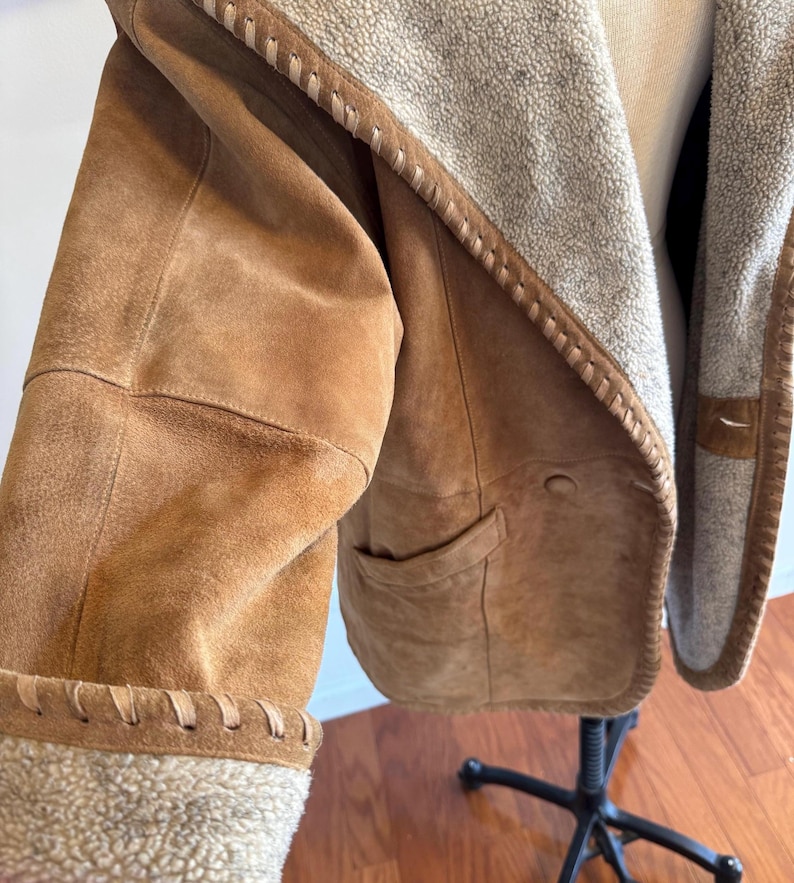 Pode incluir: Casaco de camur&ccedil;a castanho claro com forro felpudo creme e acabamento costurado. O casaco tem capuz e um bolso. As mangas s&atilde;o aparadas com o mesmo material felpudo. O casaco &eacute; exibido em um manequim.