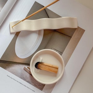 May include: White ceramic incense holder with a wavy design. A wooden incense stick is resting on the holder. The holder is on a magazine with the text "Rooster & Rice Therapy Tower Clubhouse + studio + home in Los Angeles."