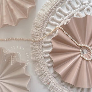 Op de afbeelding: Een set van drie beige, waaiervormige wanddecoraties met een parelketting die over een van de stukken is gelegd. De wanddecoratie is geplaatst tegen een witte achtergrond met een witte macrame cirkel met franjes.