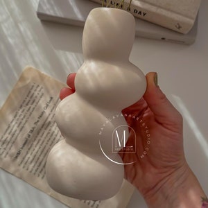 Puede incluir: Un jarrón de cerámica blanco con un diseño ondulado único. El jarrón está lleno de pequeñas flores blancas y se sostiene en una mano. El jarrón tiene el texto "MAYALAVAARTSTUDIO.COM" y "M ART STUDIO" en el lateral.