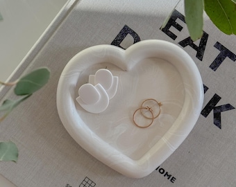May include: White heart-shaped ceramic dish with a swirl pattern. The dish is filled with two white earrings and two gold rings. The dish is sitting on a gray book with the text "EAT DRINK HOME" and "SOHO HOUSE" printed on it.