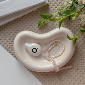 May include: A cream-coloured, kidney-shaped trinket dish with a marbled effect. The dish holds a white earbud with a black "O" logo, a pearl necklace, and a pink gemstone bracelet. A sprig of white flowers and a book are in the background.