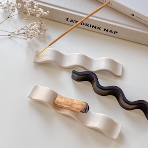 Peut inclure: Deux porte-encens en céramique blanche en forme de lignes ondulées. Un porte-encens a un bâton d'encens brun qui brûle dessus. L'autre porte-encens a un porte-encens noir sur le dessus.
