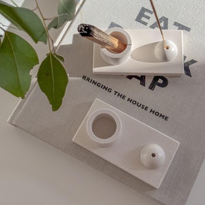 May include: White ceramic incense holder with two compartments. One compartment is for a stick incense and the other is for a cone incense. The holder is on a book with the text "Bringing the house home".