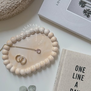 Puede incluir: Una bandeja de cerámica beige claro con un borde redondeado y un diseño texturizado en relieve. La bandeja está llena de pequeñas cuentas redondas beige. Un collar y pendientes de cristal rosa se colocan en la bandeja. La bandeja tiene el texto "MAYALAVANA ART STUDIO" escrito en ella. El texto "Aesthetic Sustainable Home Decor" también está escrito en la bandeja.