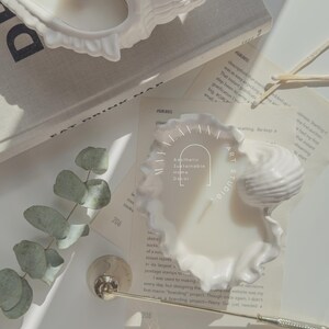 May include: White ceramic seashell candle holder with a white candle inside. The candle holder is on a white surface with a sprig of eucalyptus leaves and a book with the text "SOHO HOUS" visible. The text "Aesthetic Sustainable Home Decor" is visible on the candle holder.
