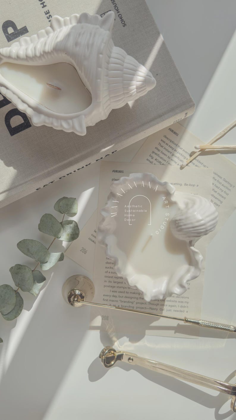 May include: White ceramic seashell candle holder with a white candle inside. The candle holder is on a white surface with a sprig of eucalyptus leaves and a book with the text "SOHO HOUS" visible. The text "Aesthetic Sustainable Home Decor" is visible on the candle holder.