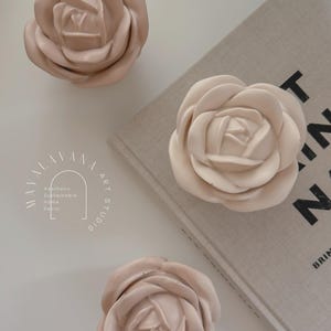 May include: Three pale pink ceramic rose-shaped drawer pulls. The pulls are arranged on a white surface with a beige book in the background. The book has the text "PRINT" in black letters on the cover.