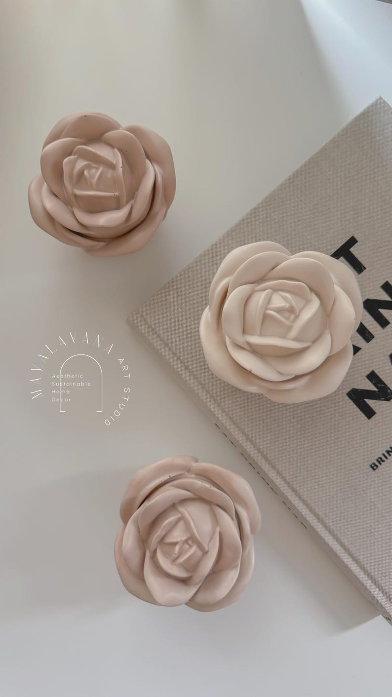 May include: Three pale pink ceramic rose-shaped drawer pulls. The pulls are arranged on a white surface with a beige book in the background. The book has the text "PRINT" in black letters on the cover.
