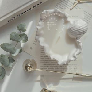 May include: A white ceramic candle holder shaped like a seashell with a white candle inside. The candle holder has a ruffled edge and a small seashell on top. The text "Aesthetic Sustainable Home Decor" is written on the candle holder.