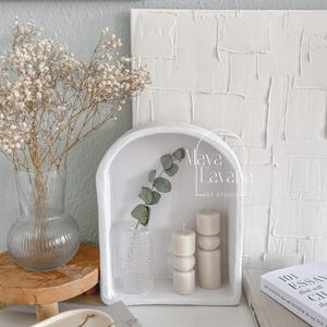 May include: White ceramic arched shelf with a small vase of eucalyptus leaves, two white candles, and the text "Maya Lavana Art Studio". The shelf is sitting on a white surface with a book and a white oval tray with a gold chain necklace.