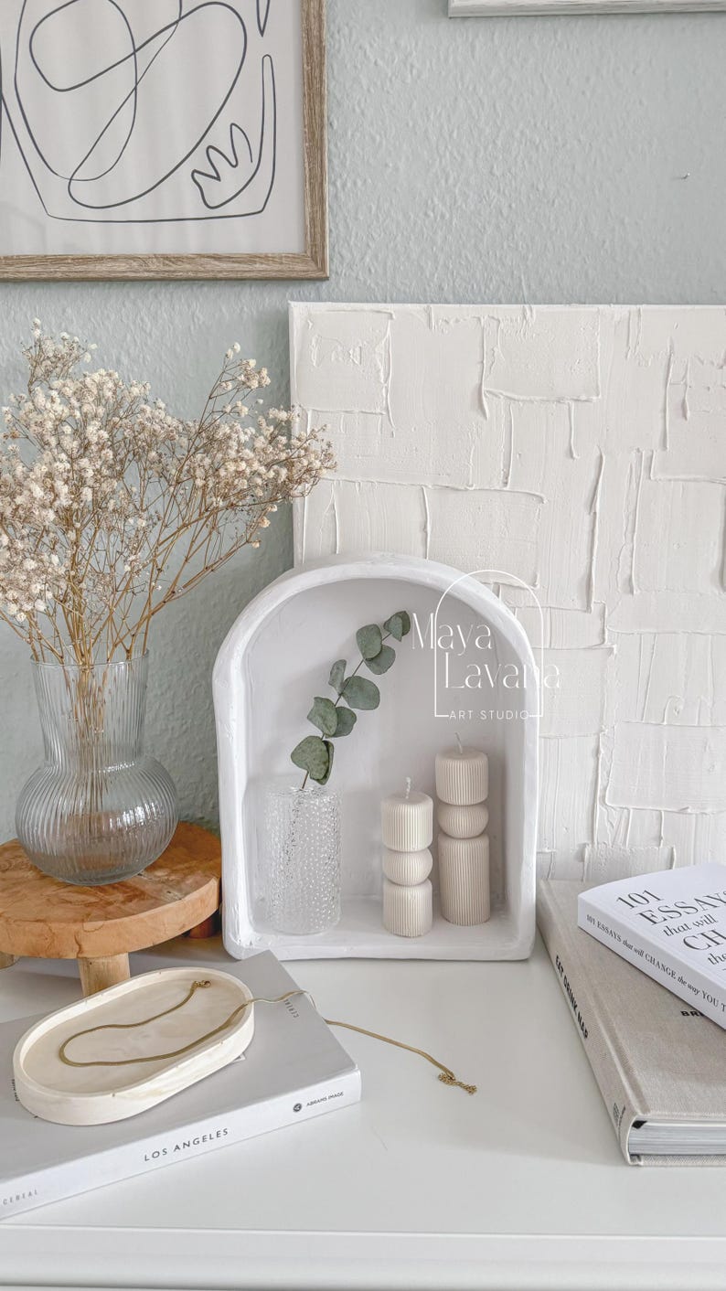May include: White ceramic arched shelf with a small vase of eucalyptus leaves, two white candles, and the text "Maya Lavana Art Studio". The shelf is sitting on a white surface with a book and a white oval tray with a gold chain necklace.