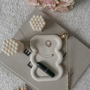 May include: White ceramic tray with a wavy edge, featuring a lipstick, a gold necklace, and two gold rings. The tray is on a book with the text "DOMO HOUSE" visible. There are two white bubble candles and a box of matches on the surface.