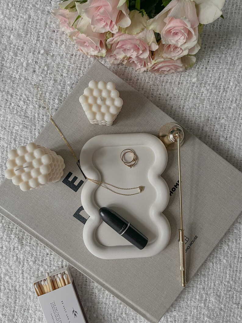 May include: White ceramic tray with a wavy edge, featuring a lipstick, a gold necklace, and two gold rings. The tray is on a book with the text "DOMO HOUSE" visible. There are two white bubble candles and a box of matches on the surface.