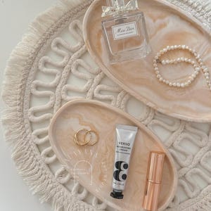 Könnte beinhalten: Zwei ovale Tabletts aus hellem braunem Harz mit marmorierten Mustern. Die Tabletts sind mit verschiedenen Gegenständen gefüllt, darunter ein Parfümflakon mit der Aufschrift "Miss Dior", eine Tube Gesichtscreme mit der Aufschrift "Verso Nourishing Cream", ein Paar goldene Creolen und eine goldene Lippenstifttube.